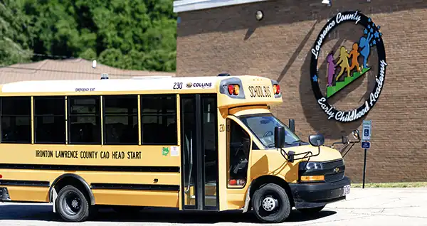 An Academy school bus parked outside the Hanging Rock Child Care building, beside the Lawrence County Early Childhood Academy logo painted on the brick wall.