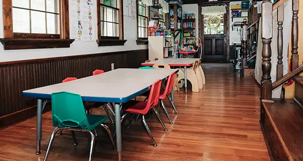 A classroom at Small Miracles Daycare, with child-sized tables and chairs on a hardwood floor beside tall windows.