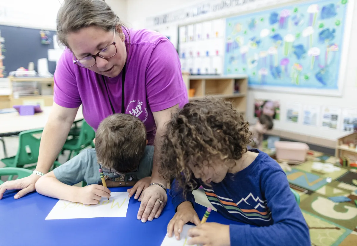 A teacher helps two children practice writing at a classroom table.