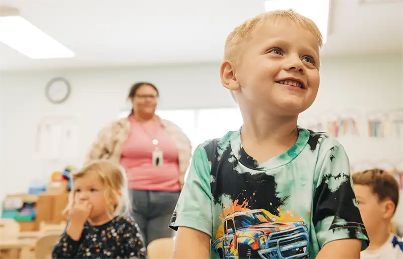 A smiling young child in a Child Care classroom with a caregiver nearby.