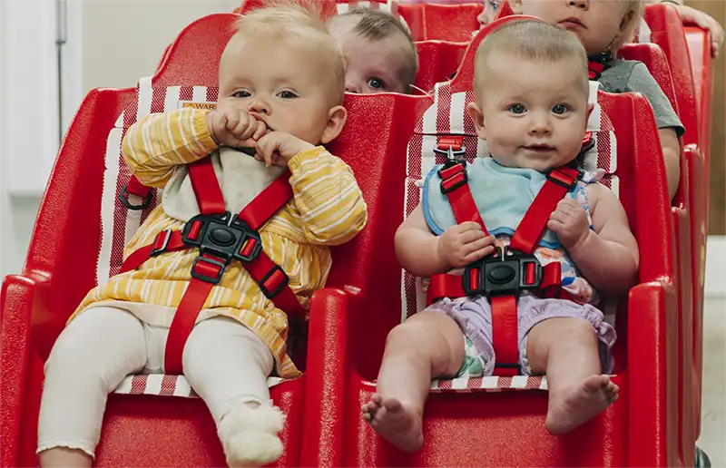 Infants buckled into red group-care seats in an Early Head Start room.