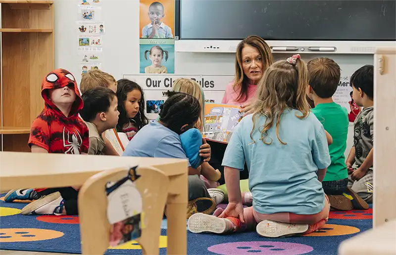 A teacher reading a picture book to preschoolers seated on the classroom carpet.