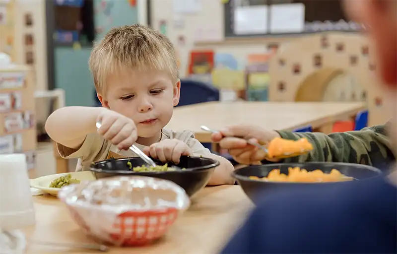 A young child eating a healthy lunch of vegetables and pasta at a classroom table
