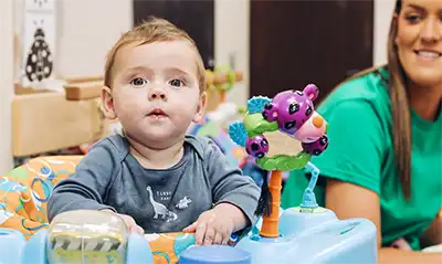 An infant exploring toys in an activity seat with a caregiver nearby